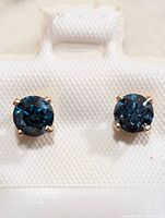 Front view of both earrings in white holder showing round blue gemstones in four-prong gold settings