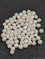 Approximately 70-80 loose natural white to cream colored freshwater pearls on a black background, showing varied sizes and near-round shapes.