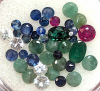 Close-up image showing a collection of various loose gemstones including green cabochons, blue sapphires, red rubies, and clear diamonds of different sizes and cuts on a white background.