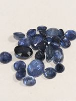 Close-up photo showing multiple loose genuine sapphire gemstones of varying shapes and sizes, deep blue color, displayed on a light surface.