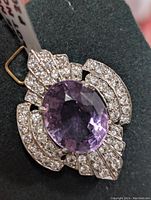 Close-up image of a sterling silver pendant with an oval amethyst stone and pavé set cubic zirconia stones arranged symmetrically around it, placed on a dark fabric background.