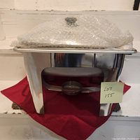 Front view of stainless steel chafing dish with glass lid wrapped in bubble wrap, sitting on red cloth.