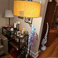 Arc floor lamp standing next to a table with various items, showing the lamp base, arm, and beige fabric shade with Lot 109 tag visible.