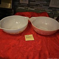 Two vintage glass mixing bowls with handles, one pink ombre with white floral berry design, the other white with geometric snowflake pattern, placed on red textured cloth.