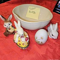Five bunny-themed items arranged on a red cloth including three bunny figurines, one decorative egg, and a cream-colored bowl.