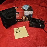 Bushnell PowerView 10x25 binoculars placed beside their original box and carrying case on a red fabric background. Includes a post-it with lot number for reference.