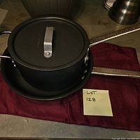 Calphalon black saucepan with lid and long metal handle on burgundy cloth next to a lot number sticky note.