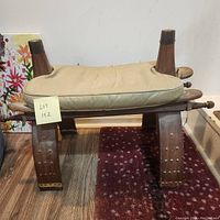 Front view of wooden camel saddle style footstool with attached beige fabric cushion showing general wear.