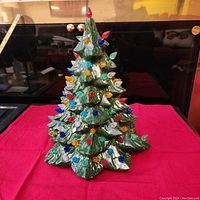 Front view of the full green ceramic Christmas tree with colorful plastic bulb ornaments attached to branches, placed on a red cloth.