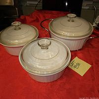 Set of three white Corning Ware casserole dishes with lids and metal handles shown on red cloth background