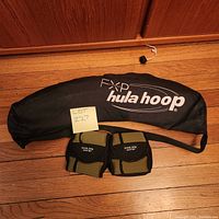 Black FXP hula hoop carrying bag with two Golden Gyre angle wrist weights in front on wooden floor
