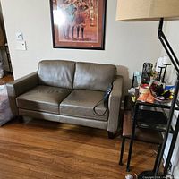 Front view of a gray leather loveseat with two seat cushions and wooden legs placed on a hardwood floor with a framed picture on the wall behind it and a side table to the right.