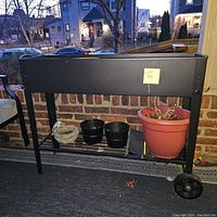 Side view of black metal box planter with three pots placed below it, including a large terracotta pot with dried plant and two smaller black pots.