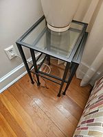 Metal nesting tables shown stacked; black metal frames with clear glass tops, decorative twisted metal ring near legs.