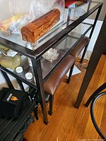 Photo showing the metal frame sofa table with glass top and lower glass shelf, one brown leather seat pushed under the table, and various small decorative items on the table.