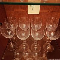 Eight clear Mikasa stemware wine glasses arranged in two rows on a wooden shelf, showing their classic rounded bowl, slender stem and flat base design.