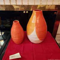 Two ceramic vases on red cloth with vintage brass fireplace in background. Larger vase with orange/white/yellow gradient next to smaller solid orange vase with texture pattern.