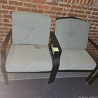 Two outdoor chairs side-by-side against a brick wall with grey cushions and black metal frames. Minor water marks visible on cushions.