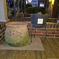 Two planters on a porch: large ceramic planter with green and floral design and black plastic planter on metal stand.