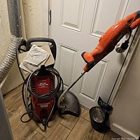 Photo showing red power washer labeled 1600 with attached hose and cables beside an orange electric grass weed wacker with black protective guard and handle, placed on a wood floor against a white door.