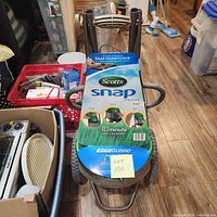 Front angle of the Scotts Snap Spreader showing product labeling, the handle system, and wheel setup on wooden floor.