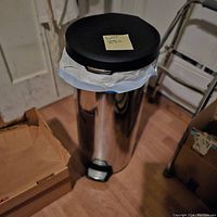 Tall cylindrical stainless steel step trash can with a black plastic lid and a foot pedal at the base. It has a trash bag placed inside the rim.
