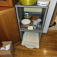Gray stackable shelf with wood grain texture, displaying ceramic bowls and cups inside, but lot includes shelves only without contents.
