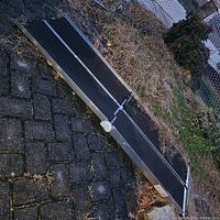 Portable metal wheelchair ramp with black textured non-slip surface and foldable feature, set outdoors on stone pavement next to grass and shrubs