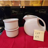 Side view showing two white BIA Cordon Bleu bowls stacked and white Fiesta Ware pitcher with handle and spout on red cloth.
