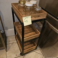 Wooden 3 shelf cart seen from an angle showing mason jars on top shelf and slatted wood design of shelves with black metal frame
