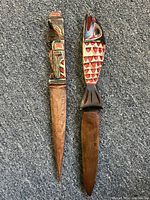 Top-down view of both letter openers side by side on a gray carpet, showing carved and painted handles and pointed wooden blades.