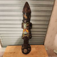 Full front view of the solid wood African mask with geometric painted details and beadwork on the face, displayed on a table in front of window blinds.