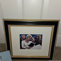 Framed artwork showing a woman reading, with colorful abstract background, in a black and gold frame with white mat.