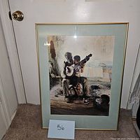 Framed artwork showing the image of a man teaching a child to play the banjo in a rustic room. The frame is gold with a light colored mat. Ragged wall and simple furniture visible in background.