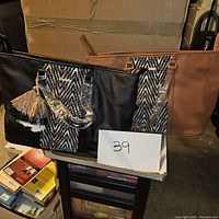 Two tote bags, one black and one brown, displayed side by side with a cardboard box behind them. Both have a central black and white geometric panel and a multi-color tassel charm on the front.