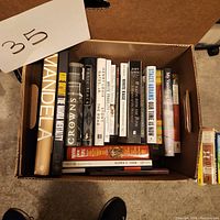 Top-down view of a box containing several used biography books with visible titles including Mandela, Kobe Bryant, Crow, Basic Black, and others.