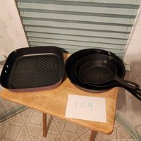 Photo of square grill non-stick pan next to two cast iron pans on wooden table with lot number 104 note