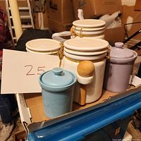 Photo showing a set of five ceramic canisters in a box, including three white canisters with clamp closures and wooden spoons, one blue canister with lid, and one lavender canister with lid.