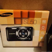 Front view of Samsung SL50 digital camera box showing camera image and key features like 10.2 MP, 27mm wide angle, 5x optical zoom.
