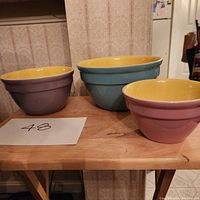 Three colorful Hall Pottery mixing bowls shown side-by-side on wooden table, colors purple, blue, and pink with yellow interiors.