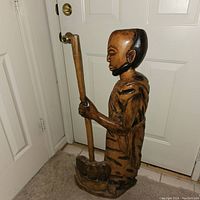 Side view of the hand carved wooden statue showing the figure holding a staff and wearing a carved garment.