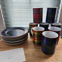 Photo of eight coffee mugs and eight plates arranged on a wooden surface showing tartan plaid patterns in multiple colors: red, blue, yellow, and green variants.