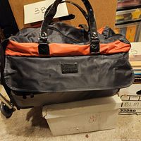 Front view of JM New York wheeled duffle bag with black patent handles, orange and gray nylon body, JM New York label on front, placed on white box.