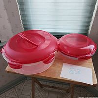 Two plastic food storage containers on a wooden table with red lids, one large with handle lid for cakes, one smaller with glass pie dish inside for pies, both labeled lot 83.