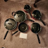 Complete view of six pots and five glass lids arranged on carpet with a card labeled 97. Pots are red with black handles and glass lids with black knobs.