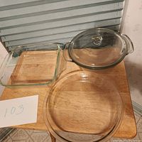 Three clear glass dishes displayed on a wooden surface: a square baking dish, a round pie dish, and a round casserole dish with lid.