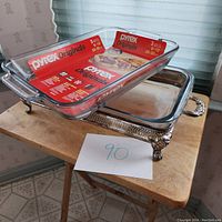 Two Pyrex 9x13 glass casserole dishes stacked on a silver-toned metal holder with decorative handles and legs, labeled with the number 90 on paper in front.