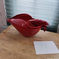 Stacked red plastic mixing and measuring bowls with handles and pouring spouts, nested inside each other on wood surface near window.