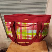 Large colorful plaid insulated bag with red handles and zipper closure on table against window blinds.