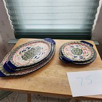 Photo showing 3 oval platters stacked on left and 2 round platters stacked on right, all ceramic with floral pattern and blue handles.